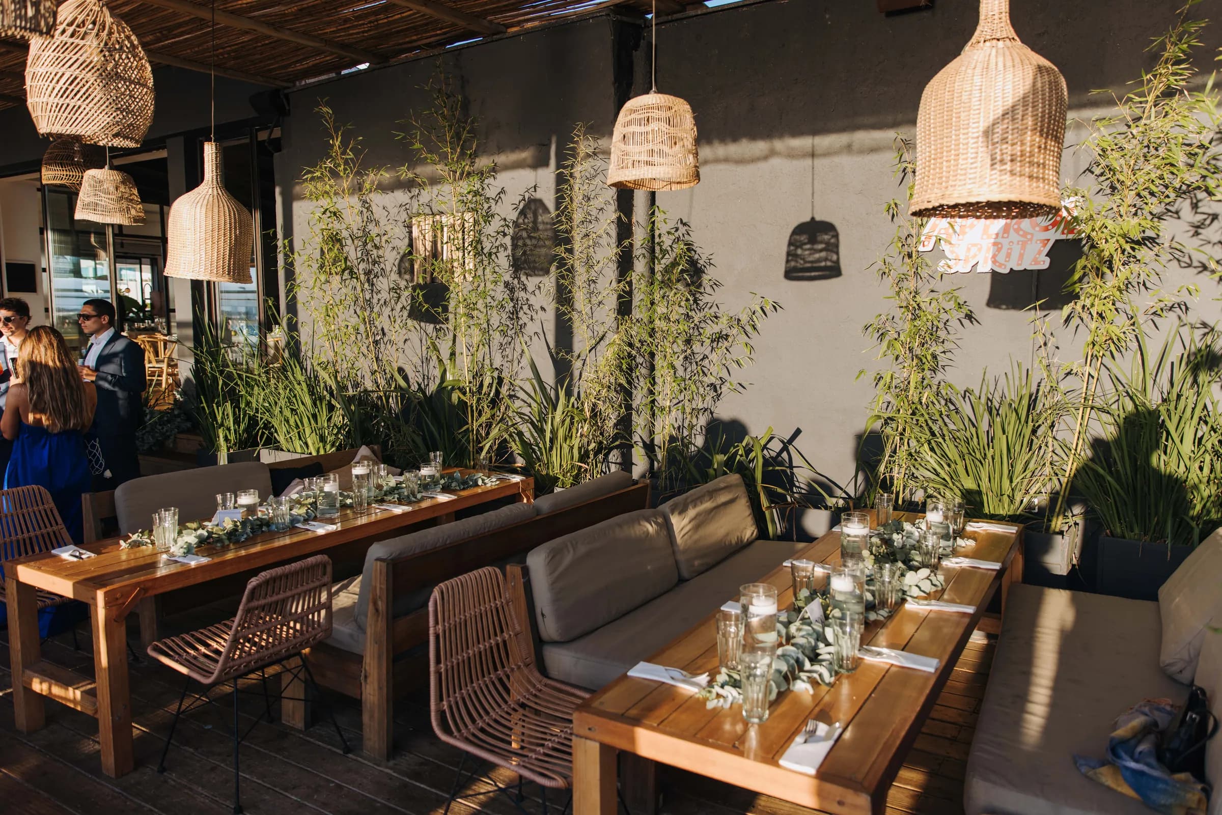 Mesas largas de madera con centros de eucalipto y velas, bajo faroles de mimbre en la terraza