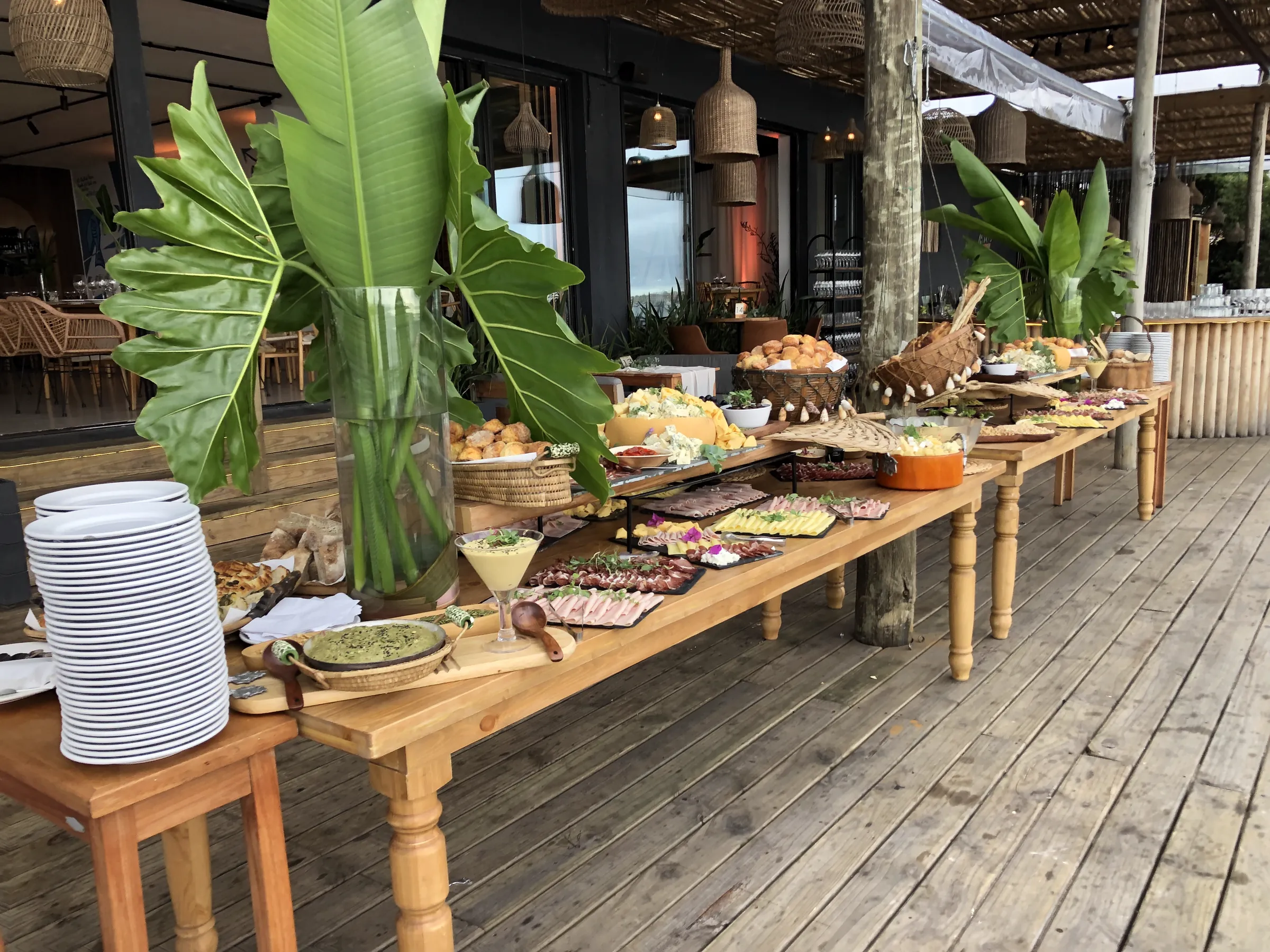 Buffet armado sobre mesas de madera en el deck de la terraza, con hojas grandes en un florero
