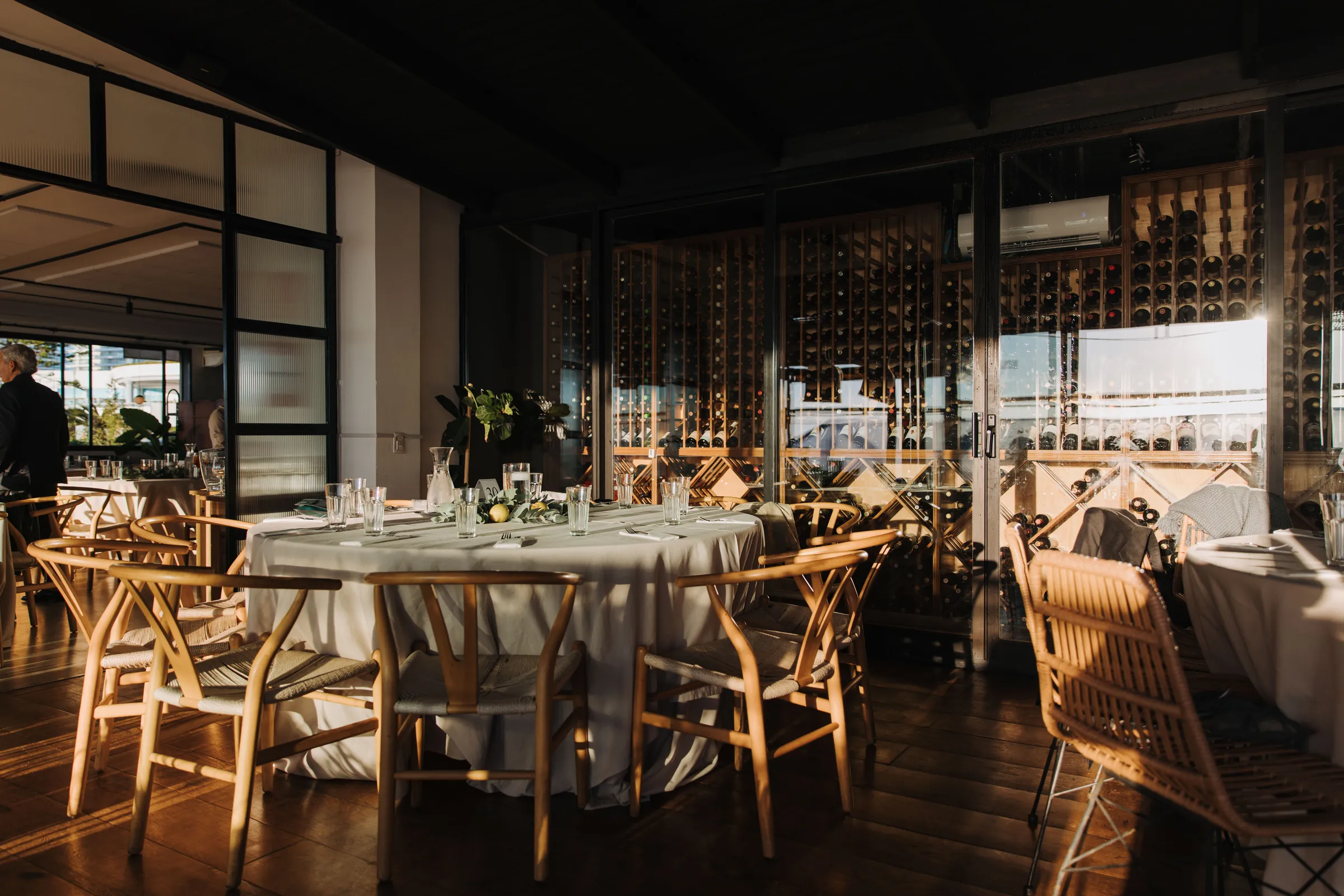 Salón con mesas redondas de mantel claro y sillas de madera, con la cava de vidrio al fondo