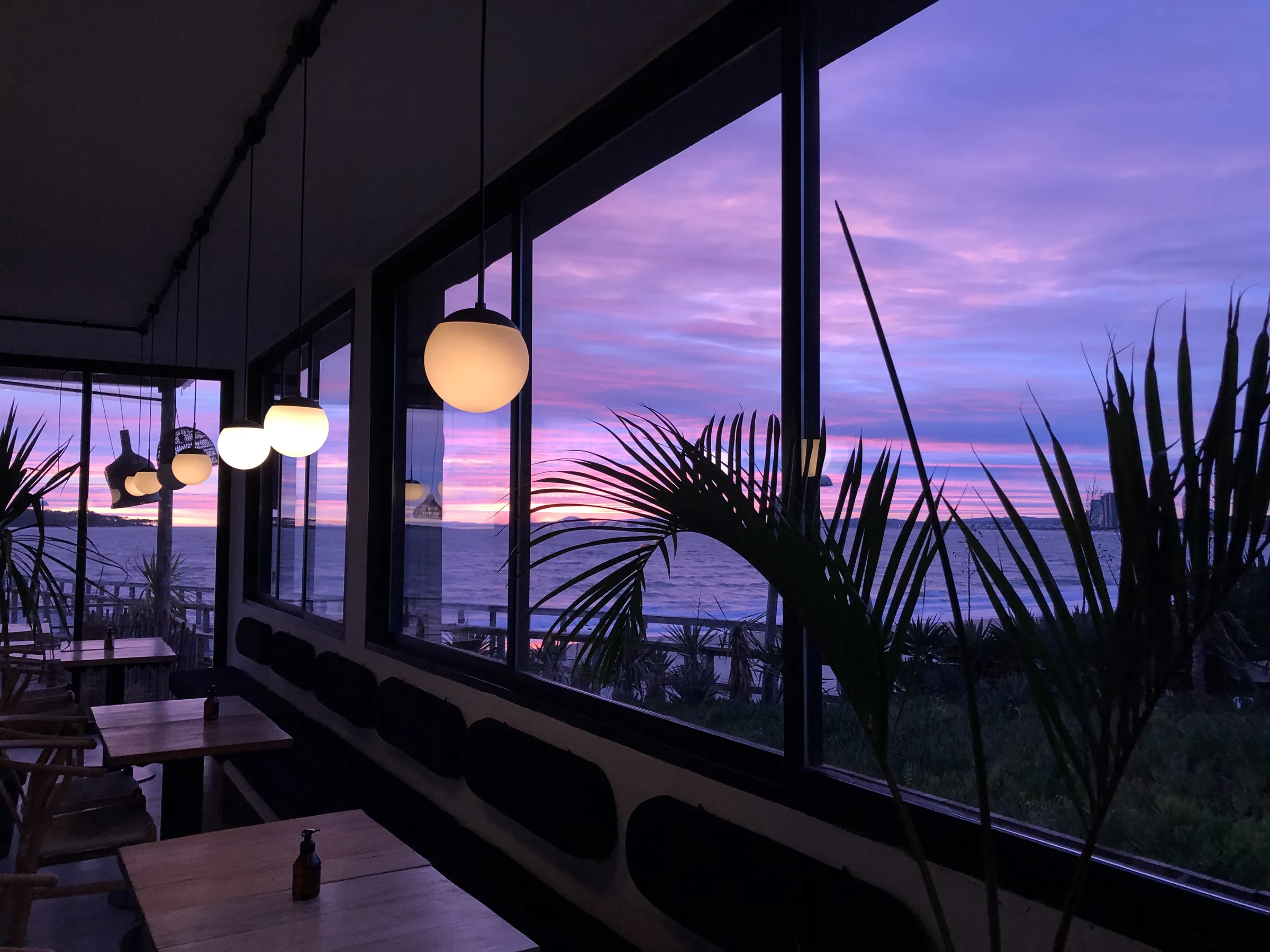Salón de Muelle 3 con ventanales, lámparas encendidas y atardecer violeta sobre el mar